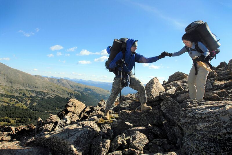 Two hikers are helping each other climb a rocky mountain. The hiker on the left is wearing a blue jacket and a large backpack, and is reaching out to help the hiker on the right. The hiker on the right is also wearing a large backpack. The sky is blue and there are some clouds in the distance. The mountain is covered in rocks and there is some vegetation in the background.